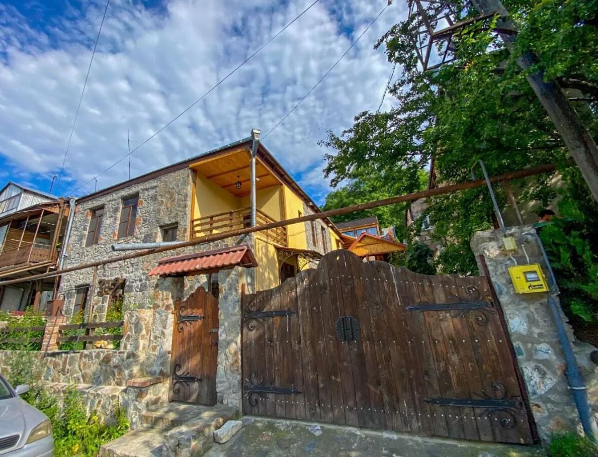 A rustic stone exterior of a house is depicted, featuring a wooden gate and a balcony above. Surrounding greenery adds to the charming atmosphere. The sky is partly cloudy, providing a natural backdrop for the cozy structure.