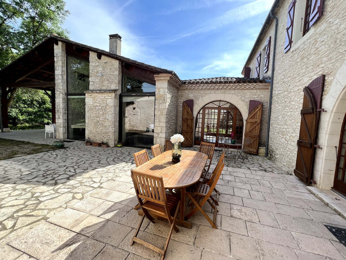 An outdoor terrace features a wooden dining table set for eight, surrounded by rustic wooden chairs. The stone walls of the house complement the serene courtyard, while large windows and doors provide a view into the inviting interior spaces.