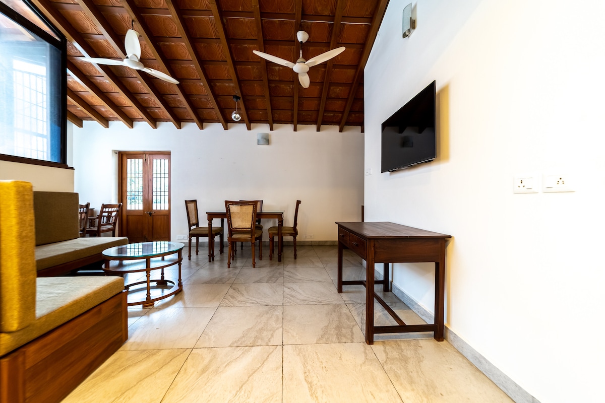 A spacious living area features a wooden ceiling and tiled flooring. A seating arrangement includes a sofa and coffee table. A dining table with chairs is positioned nearby, and a wall-mounted television is visible. Natural light enters through large windows, enhancing the atmosphere.