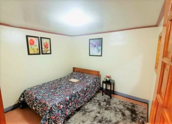 A spacious room is presented with a colorful bedspread covering a large bed. Two framed floral artworks are displayed on the walls, accompanied by a small bedside table with a vase. A plush area rug adds comfort to the wooden floor.