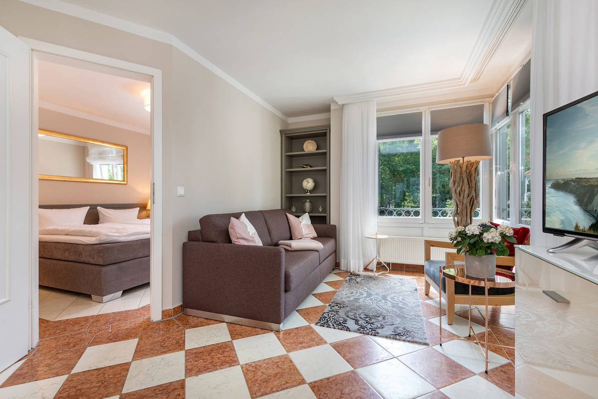 A well-appointed living area is featured, showcasing a comfortable sofa with decorative pillows. A flat-screen TV is positioned nearby, beside a stylish lamp and a small coffee table. Natural light filters through large windows adorned with light curtains, illuminating the checkerboard tiled floor.