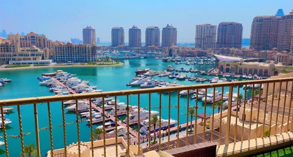 A spacious balcony offers a stunning view of the marina, showcasing numerous boats anchored in the vibrant blue water. Modern buildings line the waterfront, with a clear sky providing a bright backdrop to the serene scene.