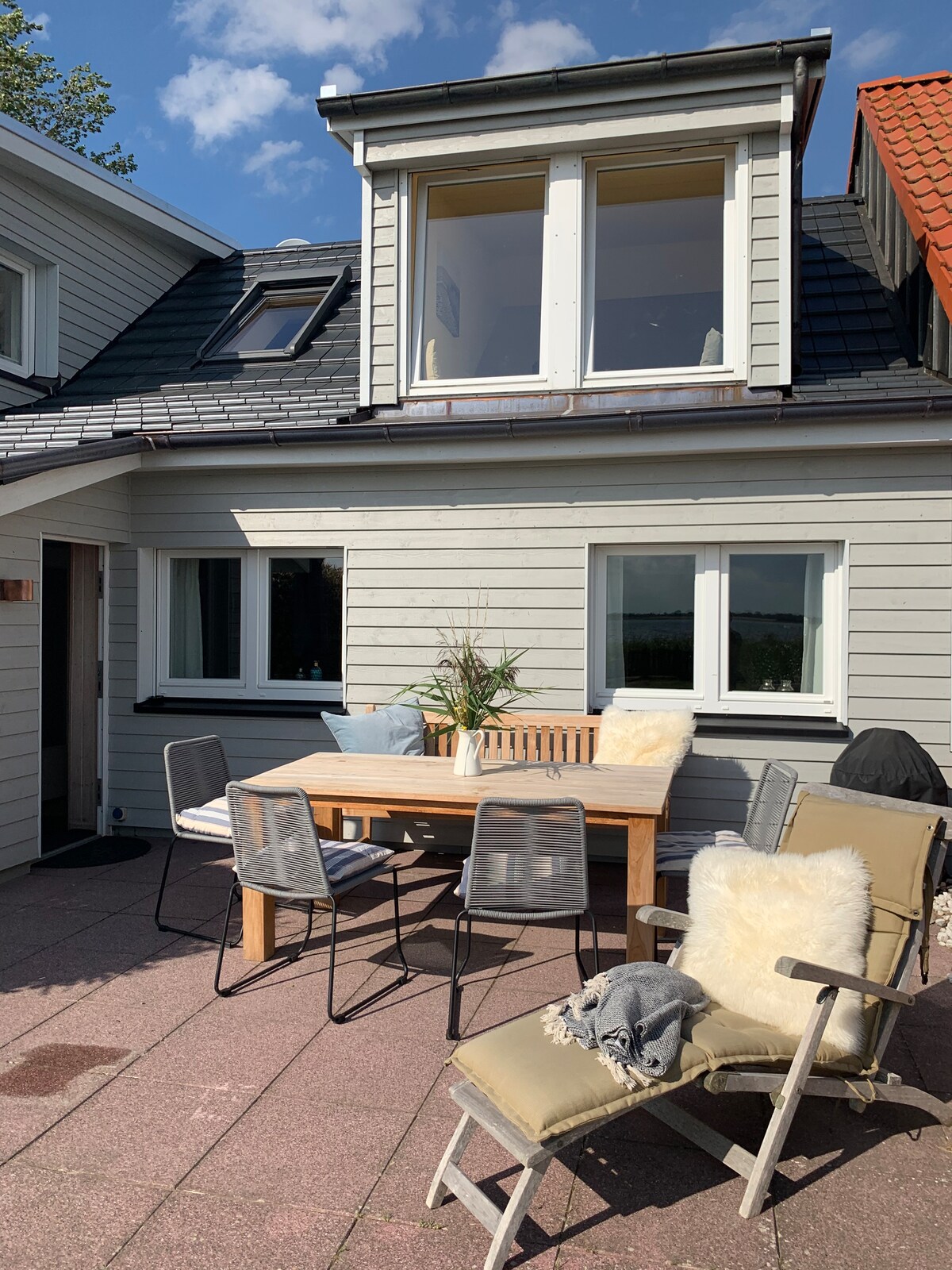 An outdoor terrace is furnished with a wooden dining table surrounded by six metal chairs. Two lounge chairs are positioned nearby, each with a soft throw. Large windows above provide natural light, while greenery adds a touch of nature to the setting.