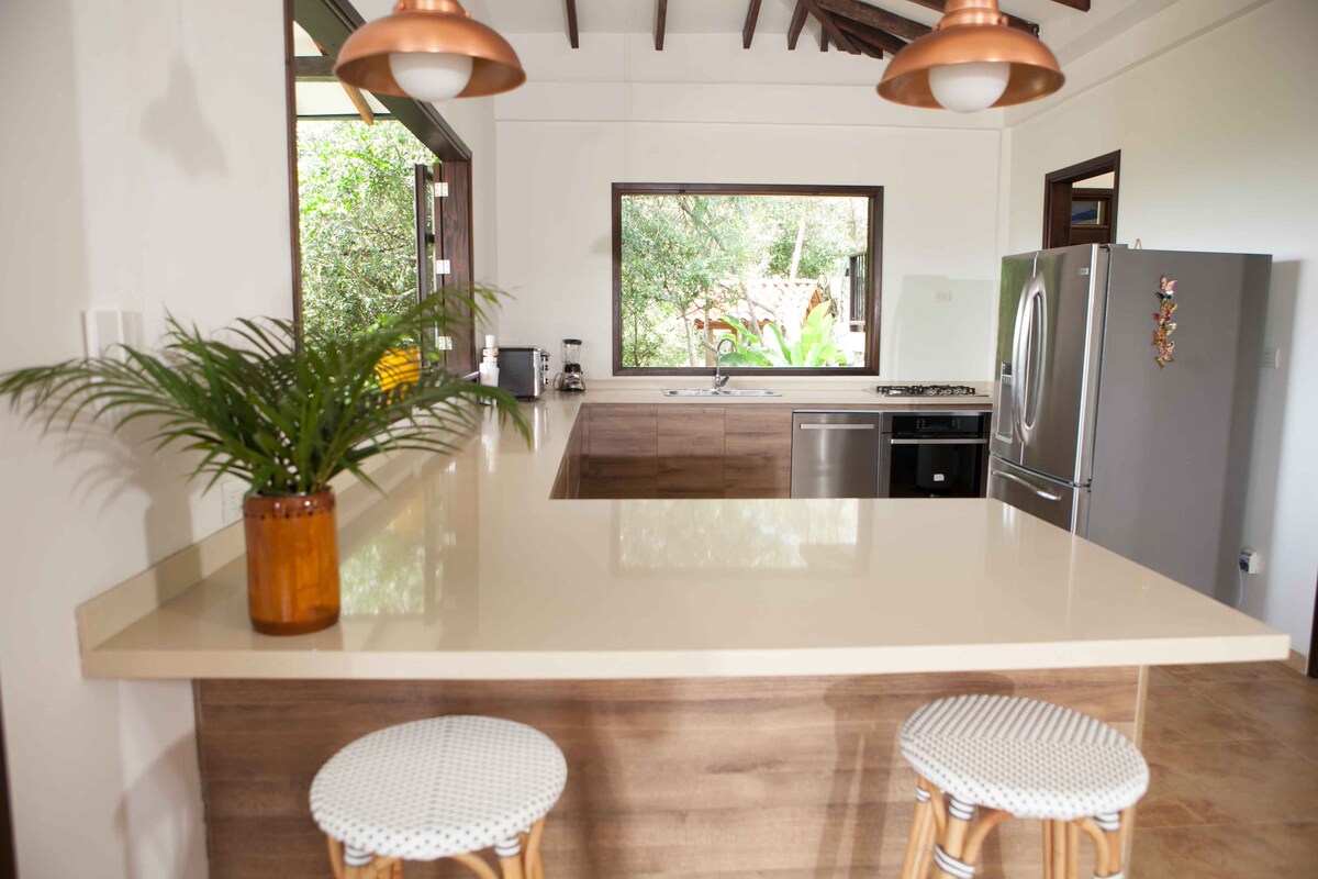 A modern kitchen space features a central island with a light-colored countertop. Two stylish bar stools are positioned nearby. Large windows allow natural light to fill the area and provide views of the greenery outside. Modern appliances are included, enhancing functionality.