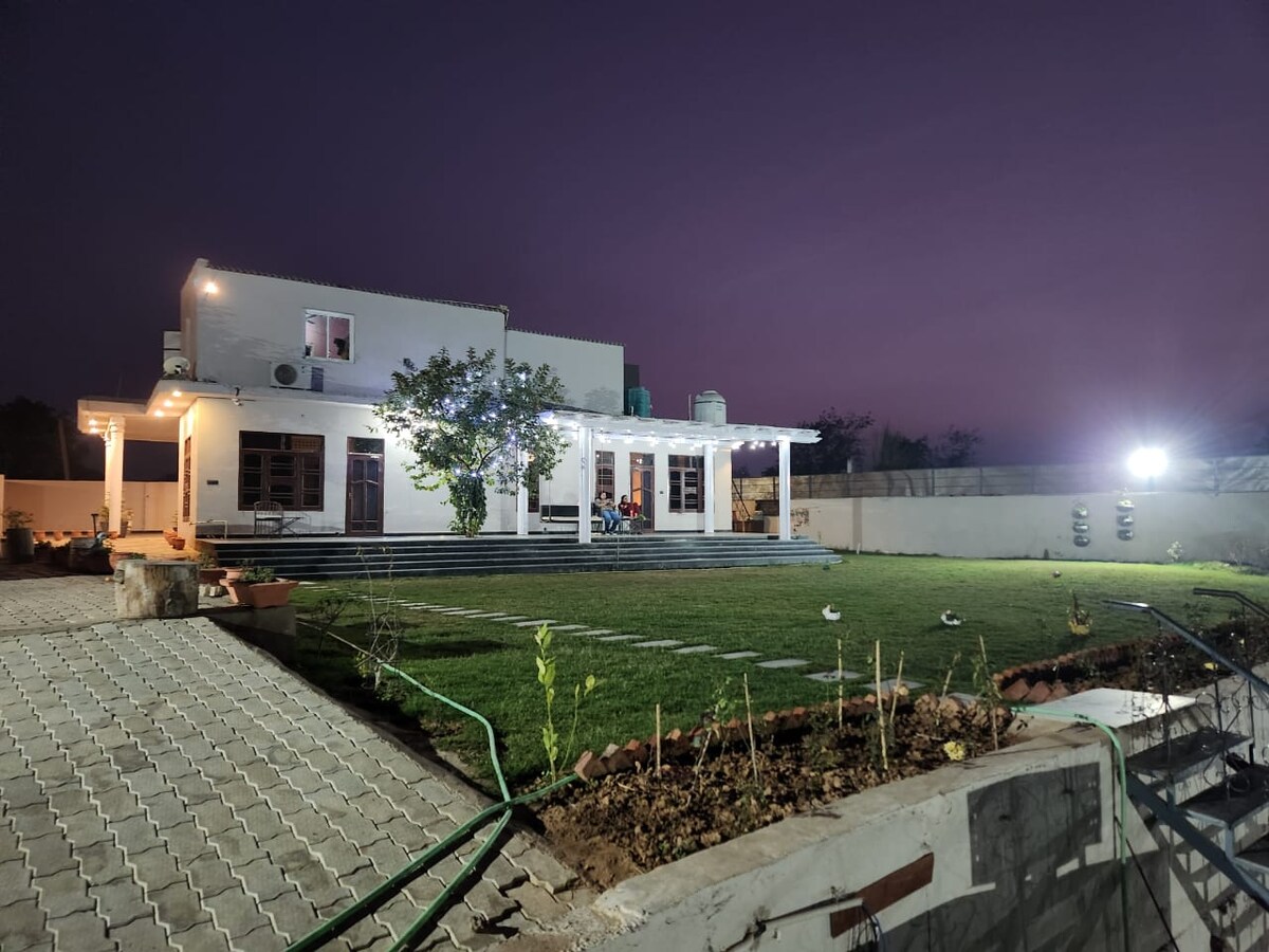 A spacious home is illuminated at twilight, showcasing a well-maintained garden with neatly arranged pathways. A large veranda with multiple seating arrangements offers a welcoming area for gatherings. Soft lighting accentuates the architectural features of the house, creating an inviting ambiance.