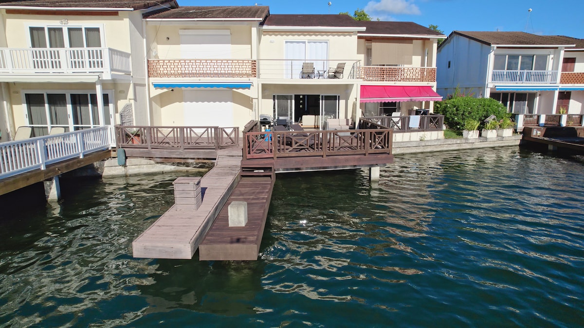 The villa is situated along the water, showcasing a wooden pier extending towards the water, providing a direct access point. The exterior features a bright facade with balconies, and various seating arrangements visible on the sun deck, creating a pleasant outdoor environment.