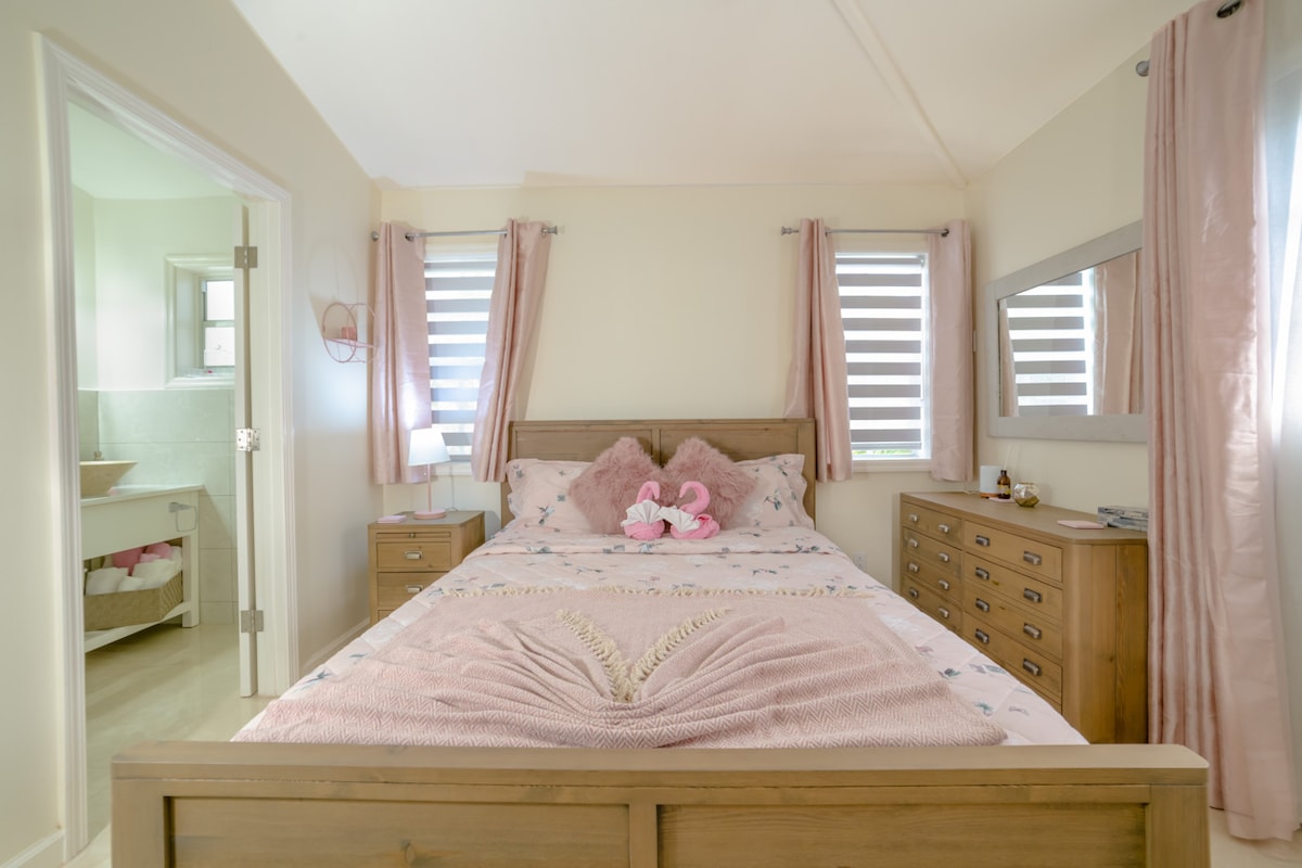 A cozy bedroom features a wooden bed with a softly designed pink bedding set. Natural light filters through slatted windows, and matching pink curtains frame the room. A dresser with a mirror and a small lamp are positioned beside the bed, providing functional space.