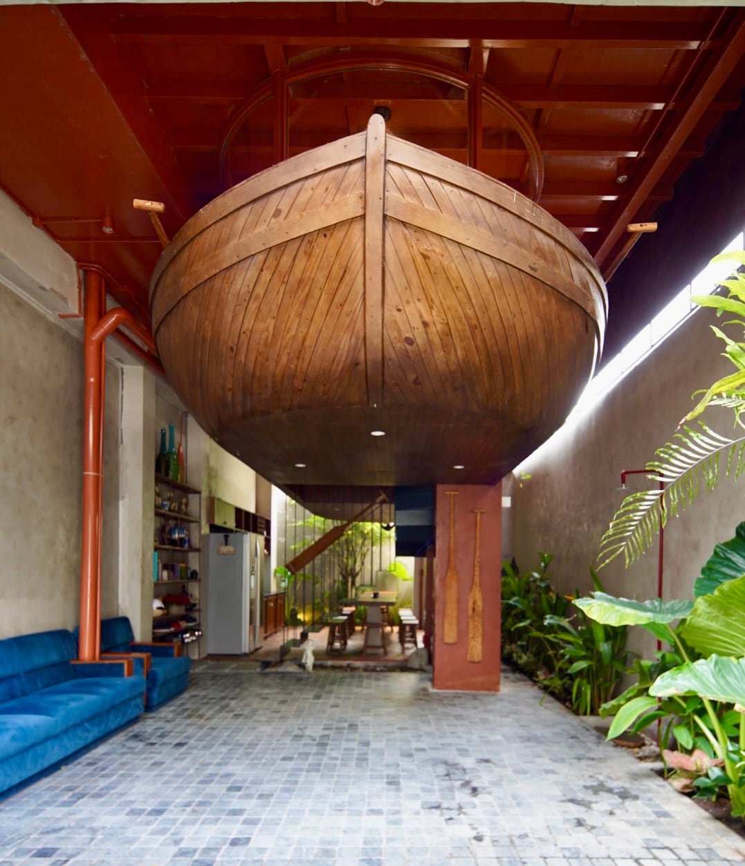 A large wooden boat is suspended from the ceiling, dominating the open living space below. The area features a blue sofa and rustic materials, surrounded by vibrant green plants. Natural light filters in through windows, creating a bright and airy atmosphere.