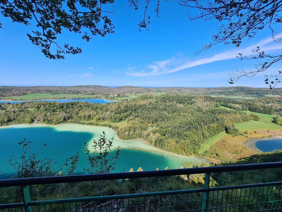A panoramic view reveals a vibrant landscape of lush green hills and sparkling lakes under a clear blue sky. The scene captures the natural beauty of the area, showcasing the tranquil waters bordered by rolling fields and dense forests.