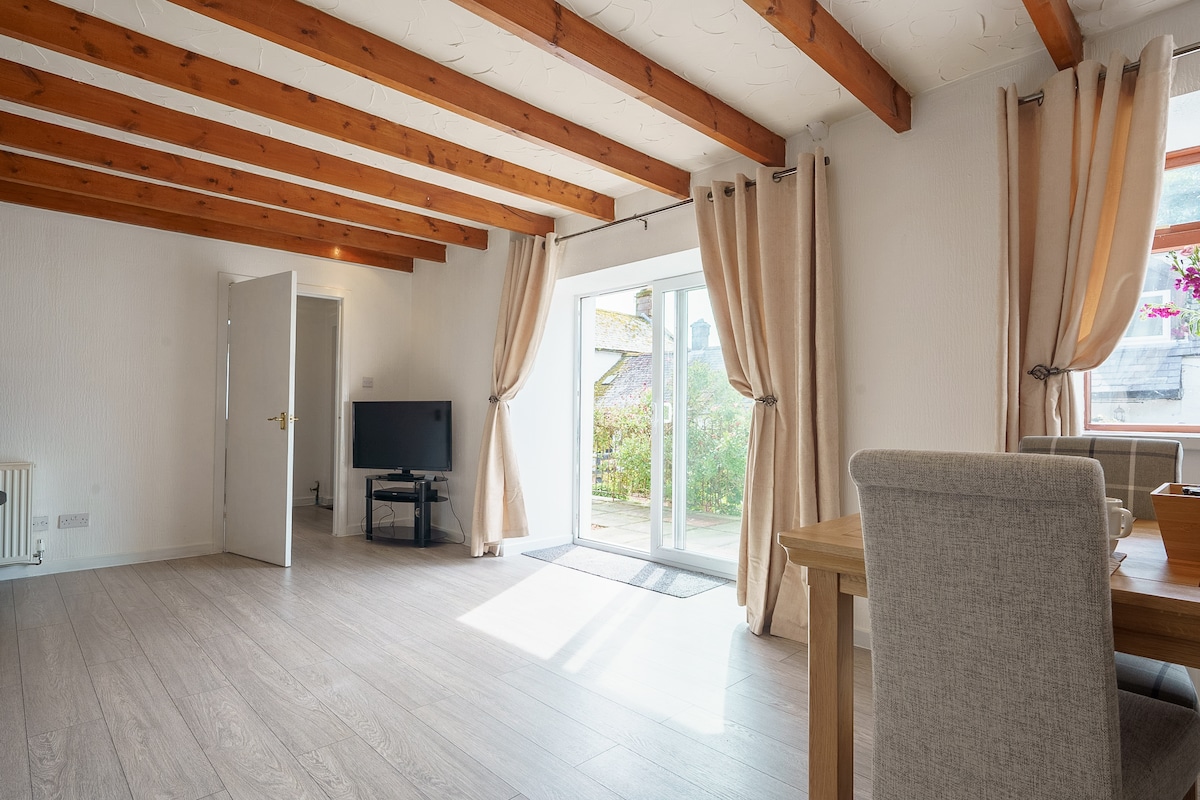 An open-plan living space showcases wooden beams across the ceiling, enhancing the airy feel. A television is positioned near a set of glass doors that lead to an outdoor area. Light-colored curtains frame the doorway, allowing natural light to fill the room.