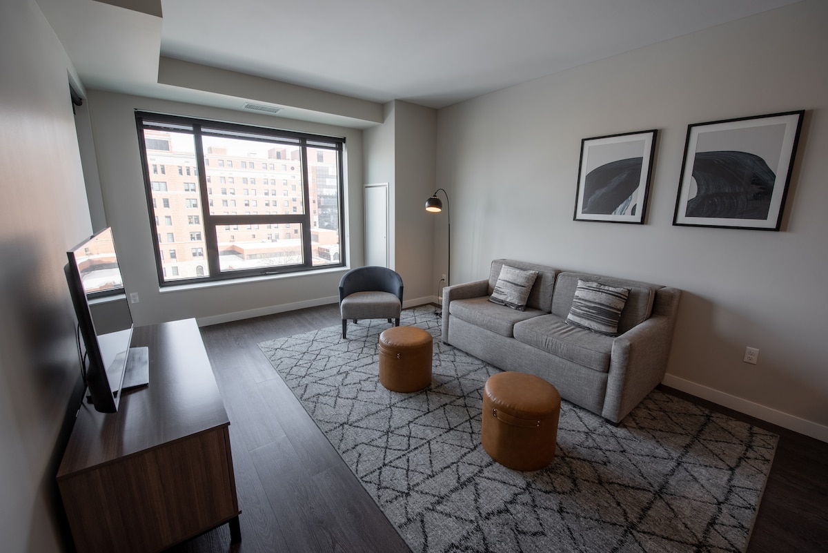 A modern living area features a grey sofa and a black accent chair, accompanied by two circular ottomans. A flat-screen television is positioned on a wooden console. Large windows allow natural light to fill the space, highlighting the neutral color palette and geometric area rug.