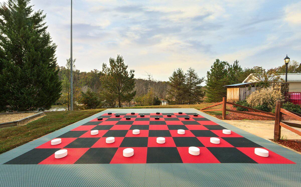 An outdoor checkerboard game area is presented, featuring a large black and red checkered pattern on a textured surface. Pieces in contrasting black and white are arranged on the board, surrounded by trees and a peaceful landscape in the background.
