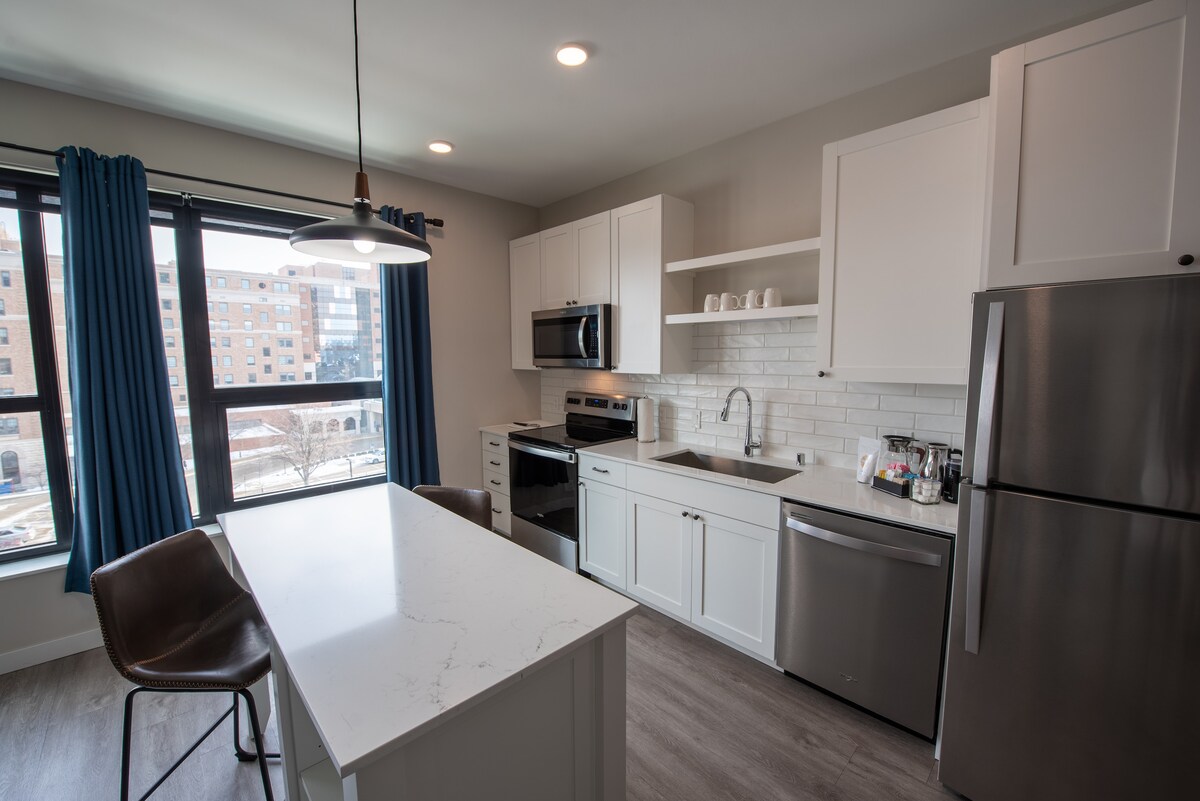 A modern kitchen is showcased with sleek white cabinetry and stainless steel appliances. A central island is positioned, providing additional counter space. Natural light floods the area through large windows, framed by blue curtains, highlighting the open and functional layout.