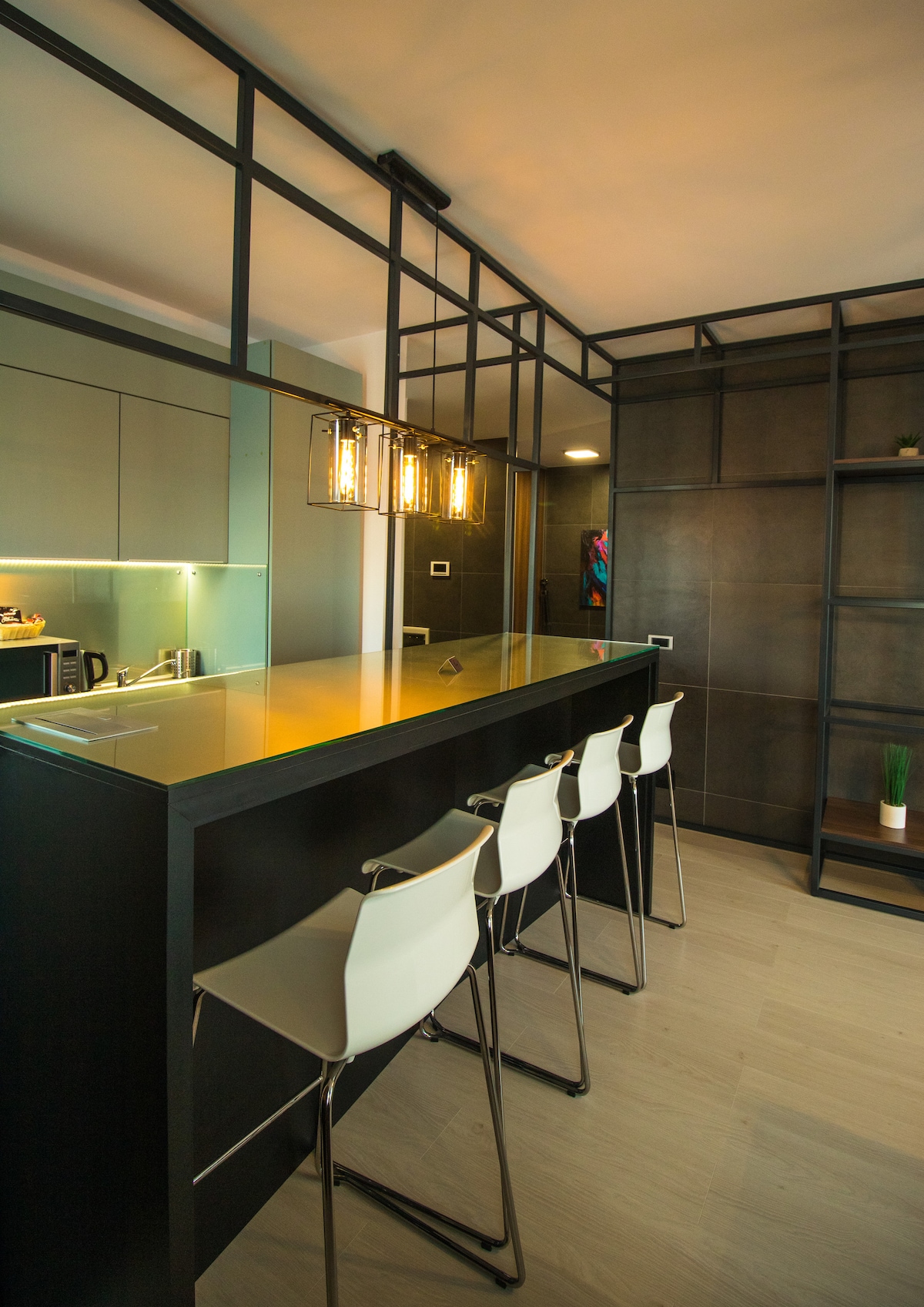 A modern kitchen area features a sleek bar with four white stools. Pendant lighting hangs above the bar, illuminating the space. Dark cabinetry provides a contemporary backdrop, while the open layout enhances accessibility and interaction with the kitchen.