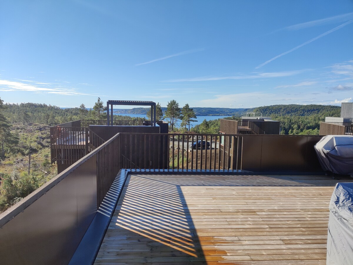 An expansive rooftop terrace is showcased, featuring wooden decking and stylish railings. Scenic views of the surrounding landscape and distant water can be observed, with clear blue skies above. Outdoor lounge furniture is positioned, inviting relaxation in this serene setting.