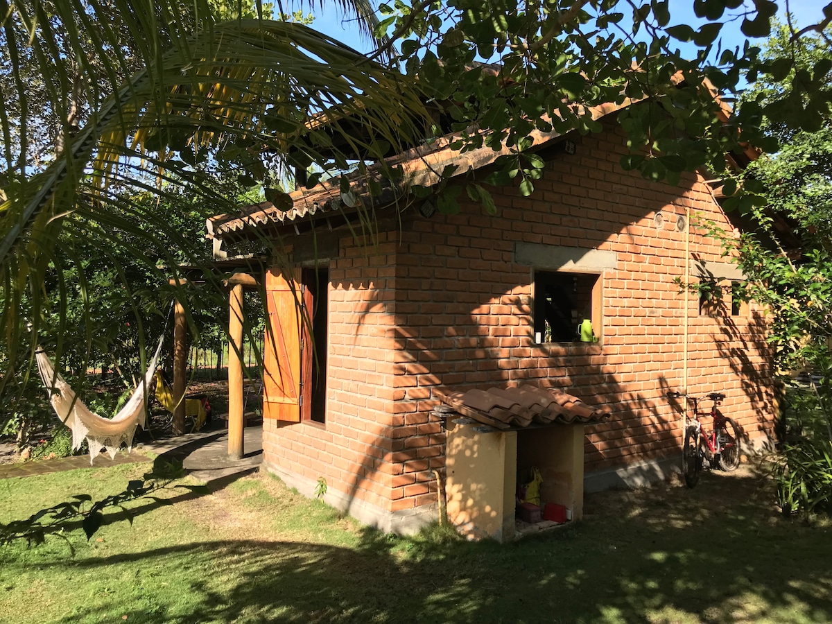 A rustic house constructed from local brick is surrounded by greenery. The structure features wooden doors and a sloped roof. A shaded area with a hammock is visible, along with a small outdoor storage space. Sunlight filters through the trees, creating a serene atmosphere.