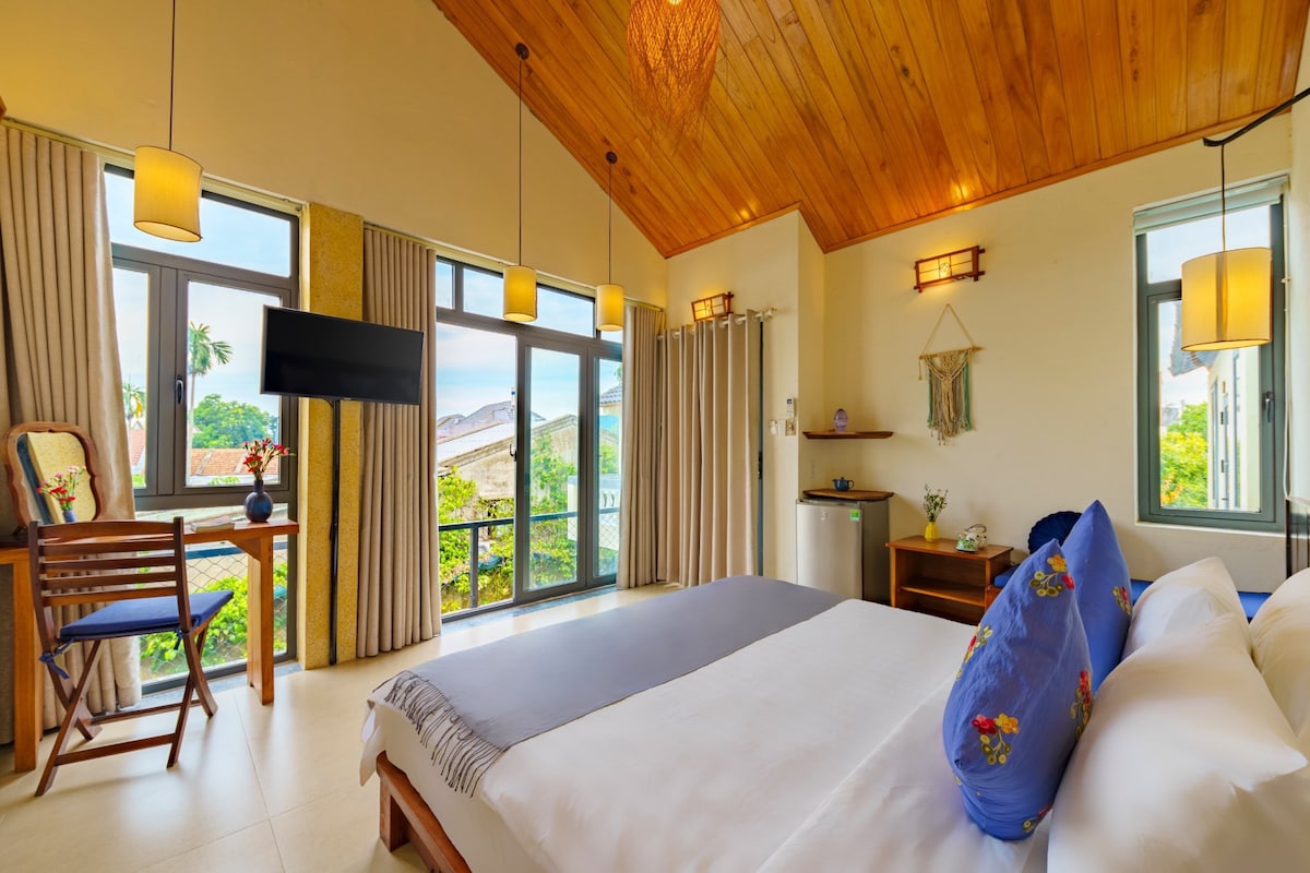 A bright bedroom features a spacious bed with a neutral-colored blanket and decorative pillows. Large windows allow natural light to fill the room, complemented by soft drapes. Furnishings include a desk with a chair and a mini fridge, enhancing the inviting atmosphere.