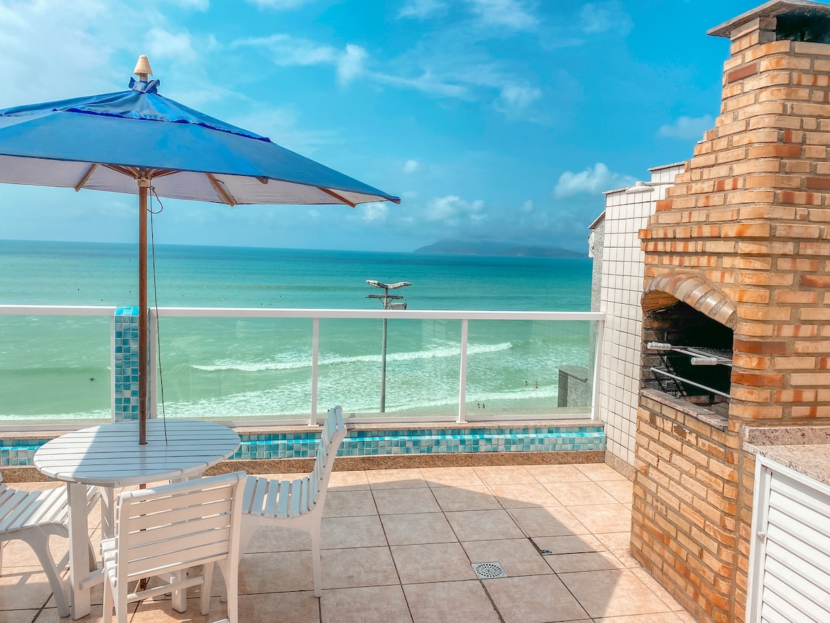 A terrace is presented with a view of the ocean, featuring a barbecue area and a dining table surrounded by white chairs. A classic blue umbrella provides shade, while the sea waves can be seen in the background under a clear sky.