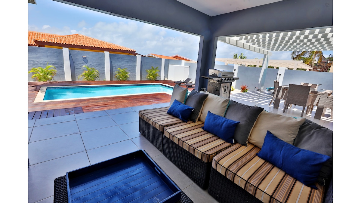 A shaded outdoor lounging area features a comfortable sectional sofa adorned with blue pillows. In the background, a pool is visible alongside wooden decking, with a dining area that includes a table and several chairs under a covered patio.
