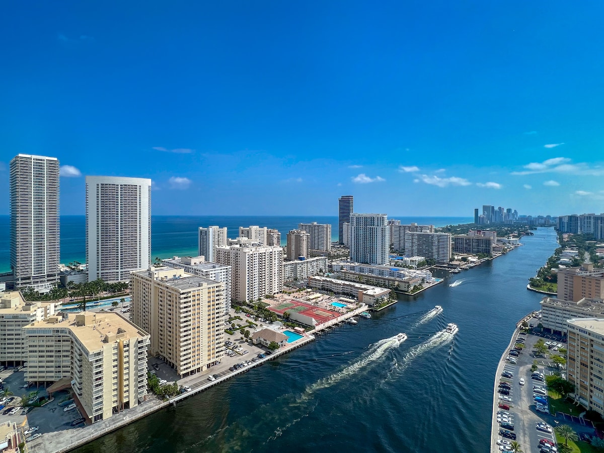 A panoramic view of the coastline features tall buildings lined along the shimmering Intracoastal Waterway. Boats glide through the water, with the Atlantic Ocean visible in the distance under a clear blue sky.