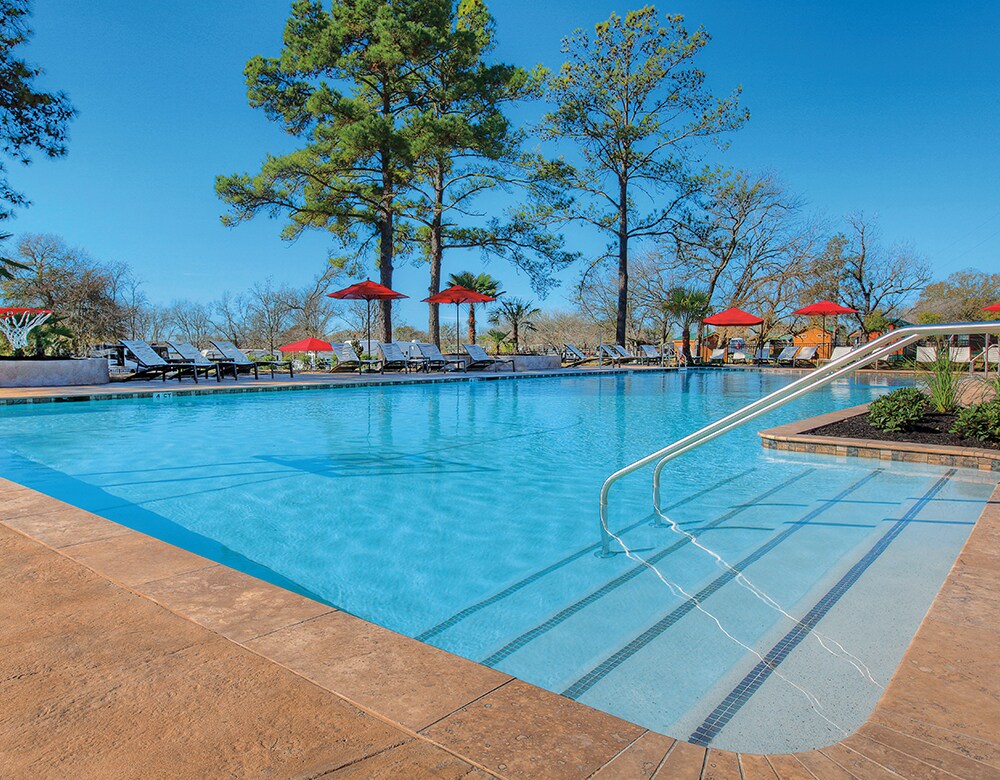 A resort-style swimming pool is framed by tall trees and lounge chairs. Red umbrellas provide shade over selected areas, while the clear blue water reflects the sky. A gradual entry is visible along one side, leading to the pool's inviting depths.