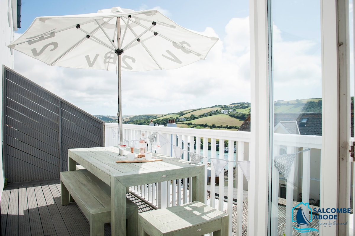 An outdoor balcony space features a wooden dining table surrounded by benches, ideal for four guests. A large umbrella provides shade, while panoramic views of the countryside are visible in the background. The setting invites relaxation and dining in a tranquil environment.