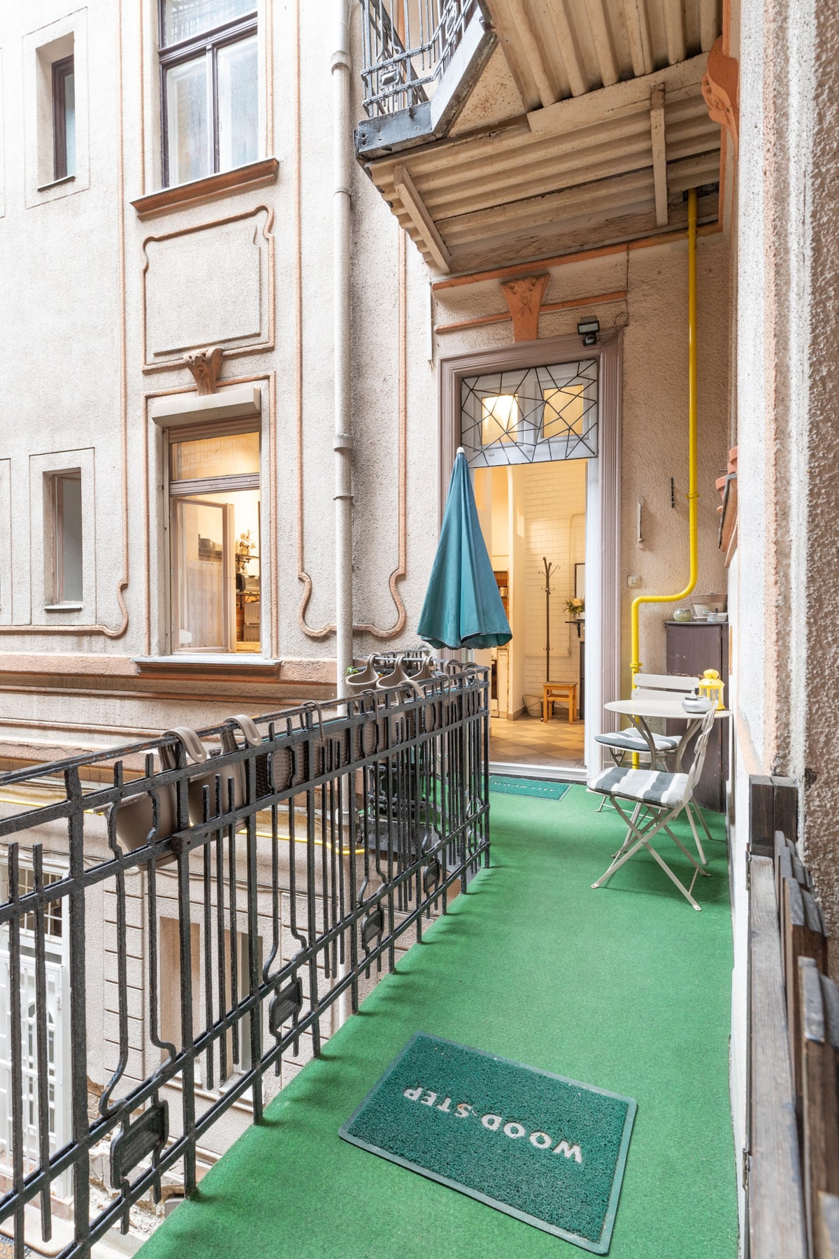 A balcony area is featured, with a green carpet underfoot and a small folding chair beside a table. An umbrella stands to provide shade. The view leads to the entrance of the cozy home, adorned with a decorative glass door.