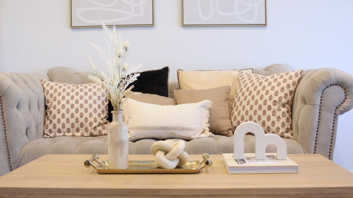 A neutral-toned living area features a tufted sofa with patterned throw pillows and a selection of decorative accents. A wooden coffee table is adorned with a tray containing a vase, a decorative knot, and a book, enhancing the space's modern aesthetic.