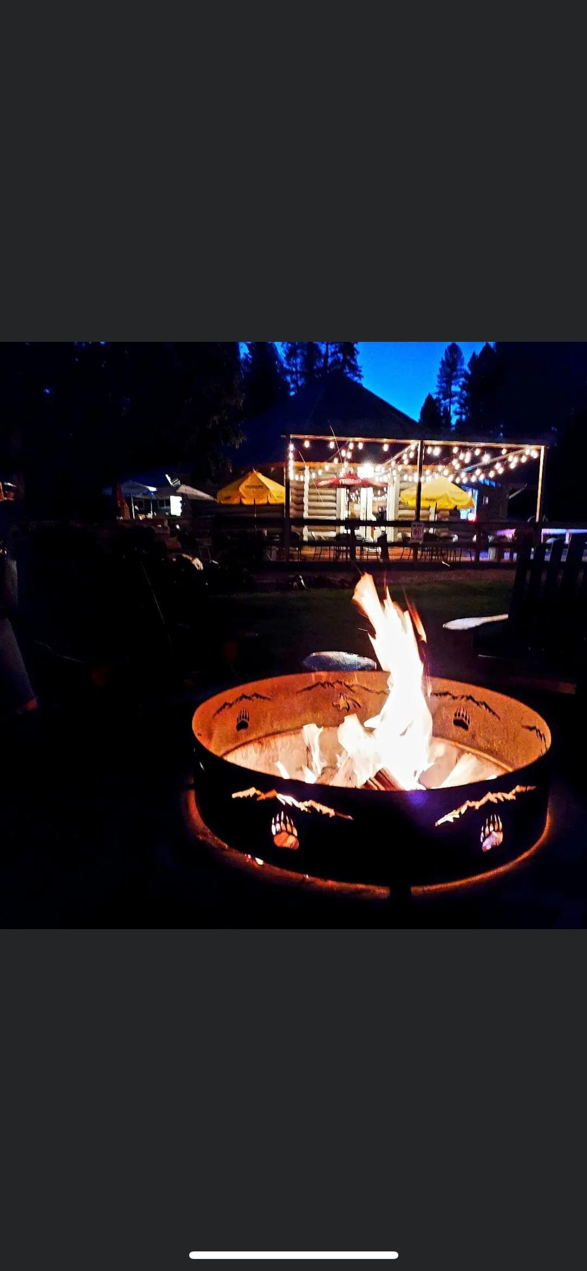 A firepit surrounded by wooden seating provides a warm glow as flames dance in the nighttime. In the background, string lights illuminate a covered patio and cabin structure, contributing to a relaxed outdoor setting.