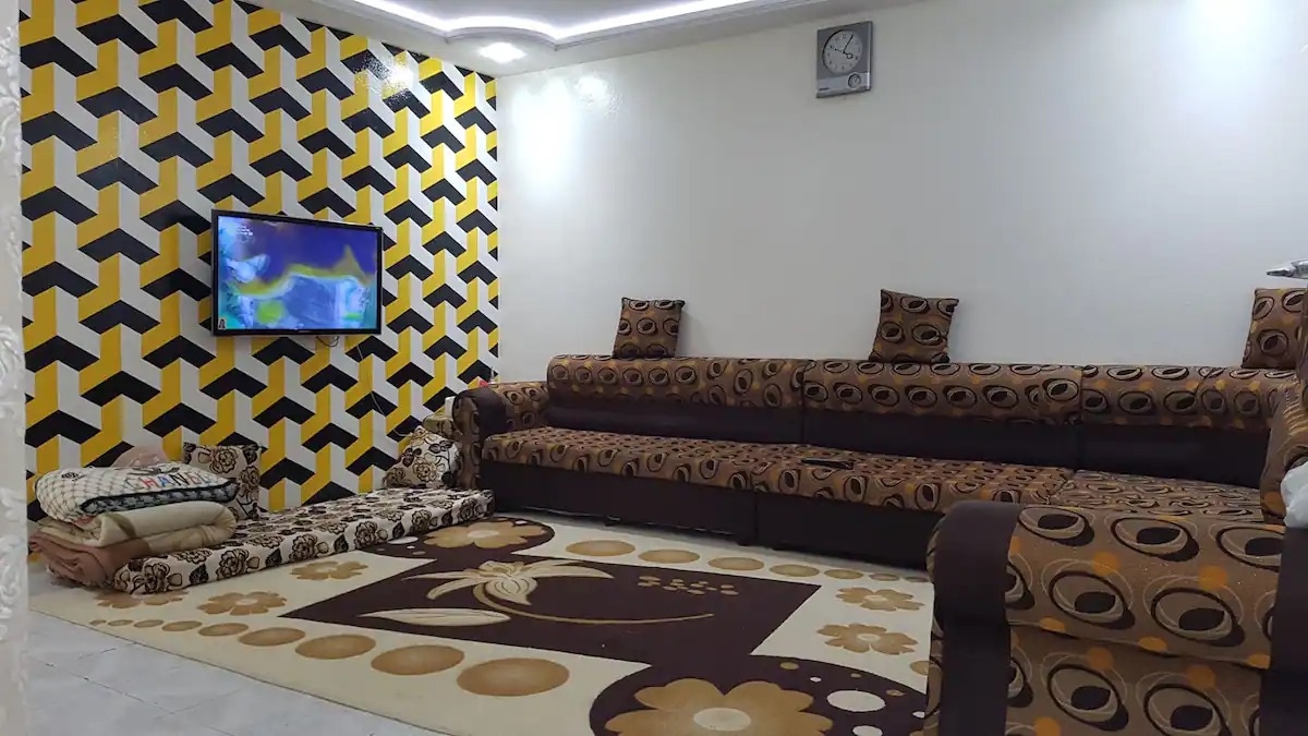 A spacious living area features a distinctive patterned wallpaper in yellow and black. A large sectional sofa in earth tones is accompanied by several decorative cushions. A flat-screen TV is mounted on the wall, while a large area rug with floral design covers the floor.
