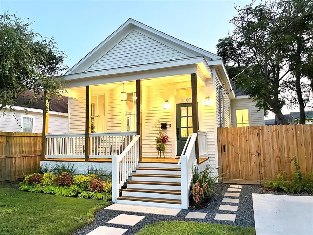 A charming bungalow exterior is presented, featuring a welcoming front porch with hanging lights. Lush greenery and neatly arranged plants line the pathways leading to the entrance. A fenced yard provides privacy, with a small driveway visible to the side.