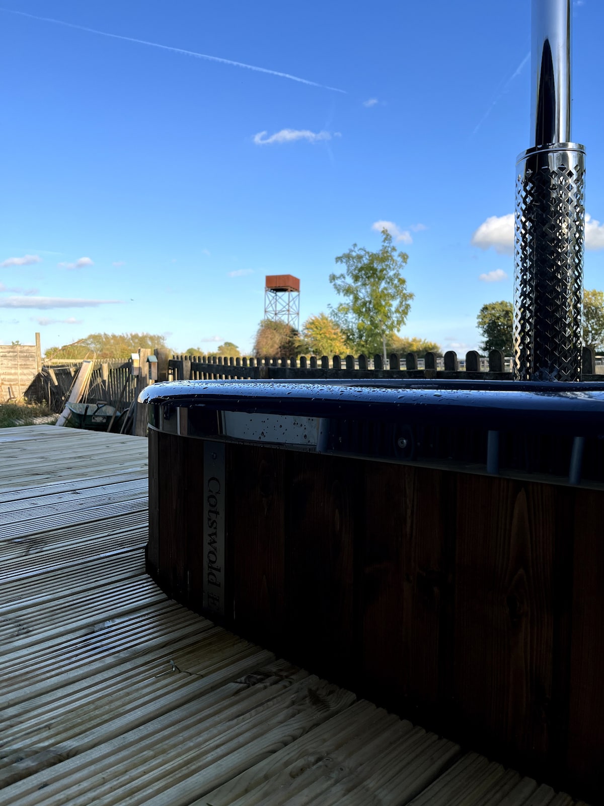 A wood-fired hot tub is positioned on a wooden deck, showcasing its rounded wooden exterior. The backdrop features clear blue skies and scattered clouds, with distant greenery and a structure visible in the distance, contributing to a rustic, serene environment.