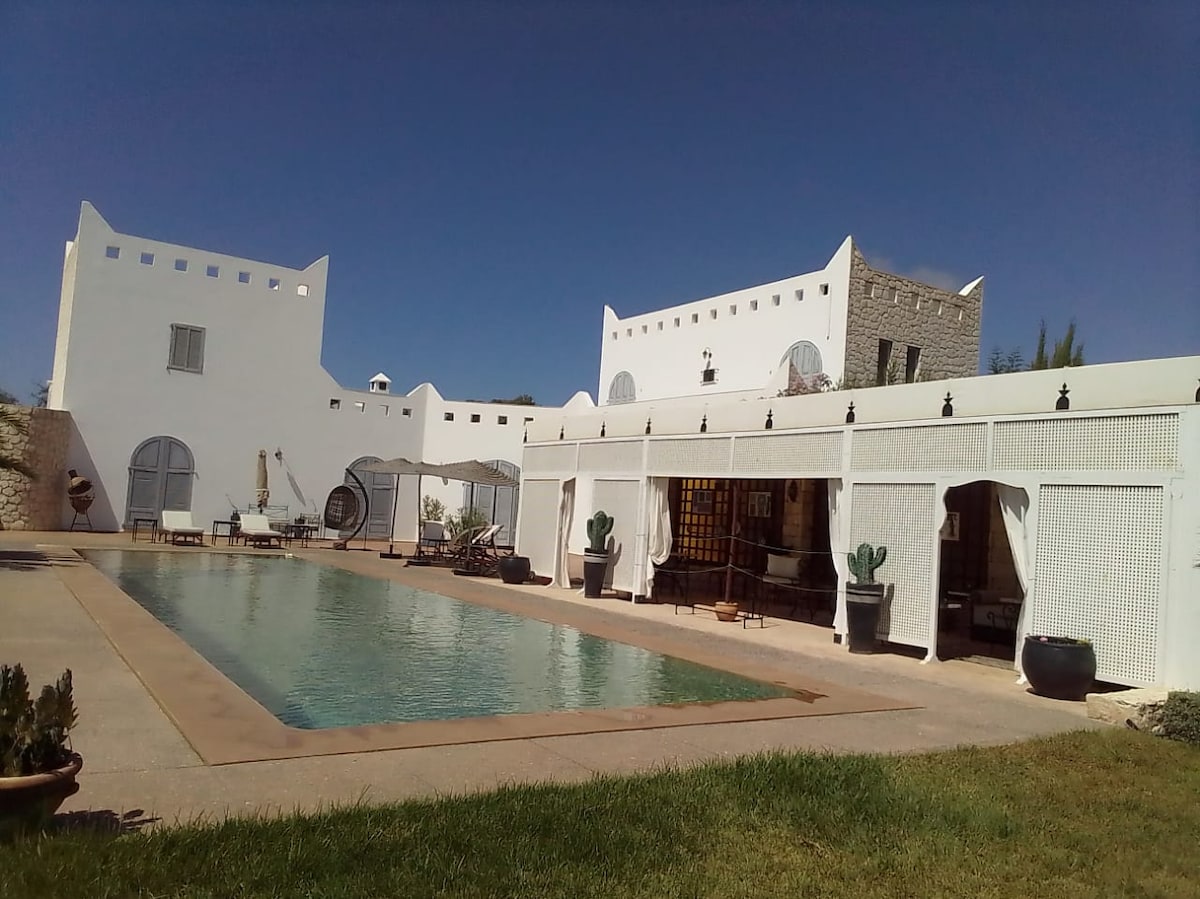 The exterior of the villa is presented, showcasing a large swimming pool surrounded by a spacious terrace. White walls define the structure, complemented by window details. Lush greenery is visible around the pool area, contributing to a serene environment.