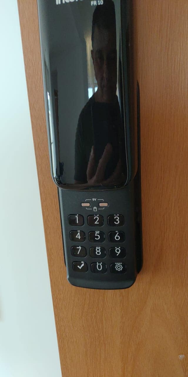 A modern digital keypad is mounted on a wooden door. The sleek black design features numbered buttons and a touchpad for code entry, offering a contemporary and secure access option.