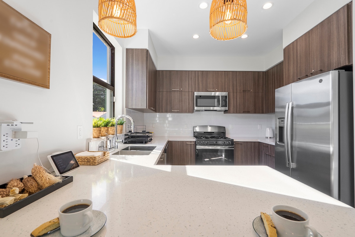 A modern kitchen is visible, featuring dark wood cabinetry and stainless steel appliances. Natural light floods the space through a large window. A kitchen island offers seating, with a glimpse of freshly brewed coffee and snacks arranged on the countertop.