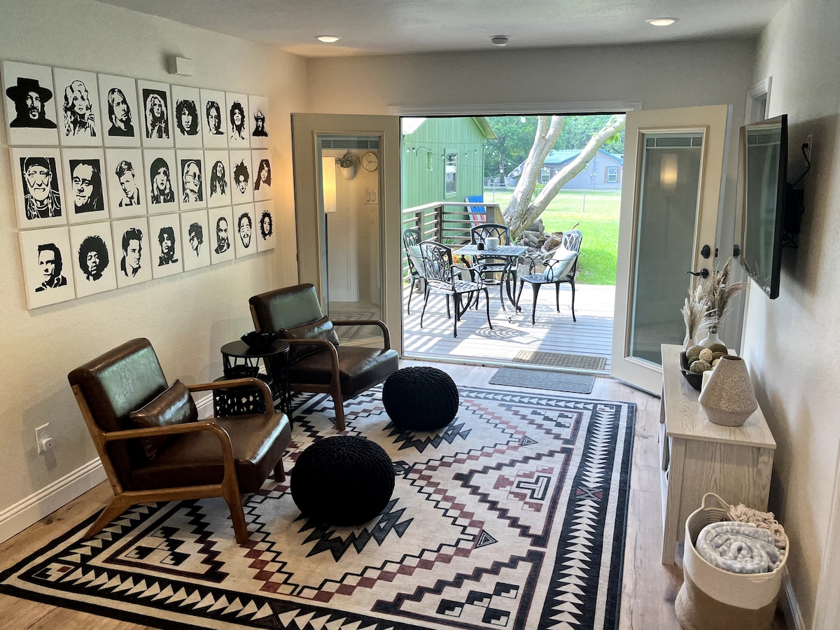 A welcoming living area is showcased, featuring two stylish chairs and a patterned rug. Large double doors open to a shaded outdoor deck area with a dining table and chairs, accessible from the inside, inviting a seamless transition between indoor and outdoor spaces.