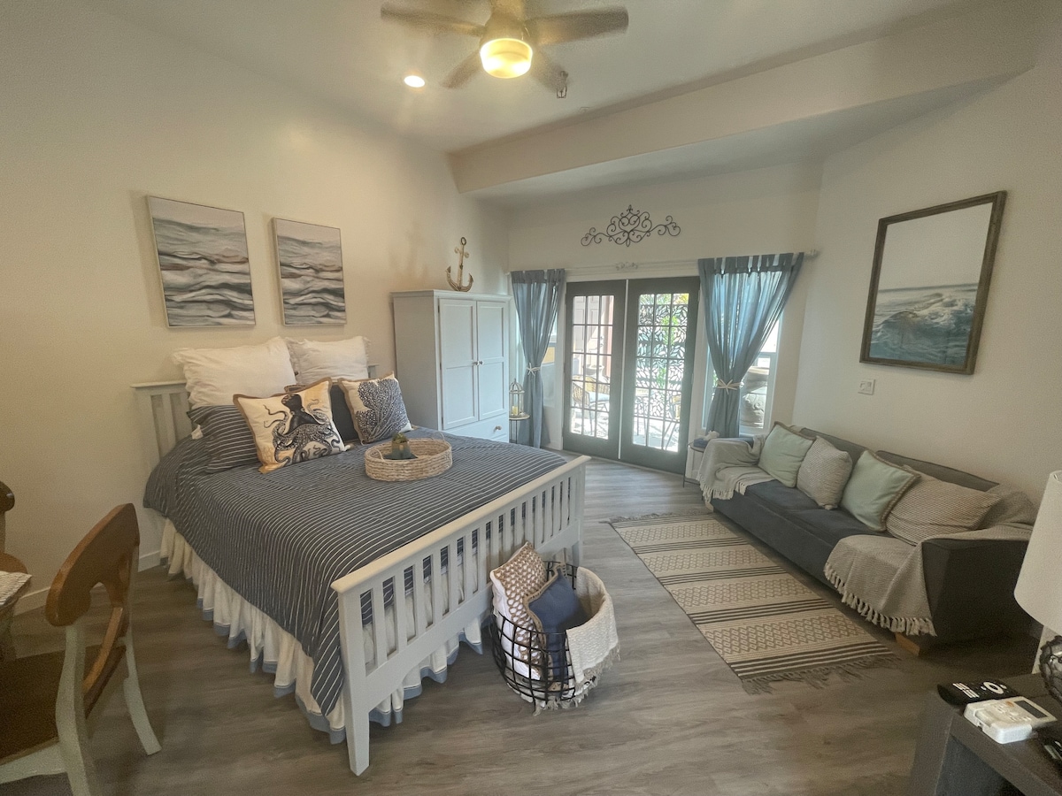 A spacious studio room features a comfortable bed with decorative pillows and a striped blanket. A cozy seating area with a dark sofa and soft cushions is present. Light streams in through glass doors that open to a private outdoor space, enhancing the airy ambiance.
