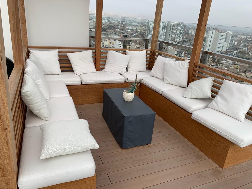 An outdoor seating area is displayed, featuring a large wooden deck surrounded by a skyline view. The space includes a sectional arrangement with white cushions and a dark fabric-covered coffee table, offering a comfortable spot to enjoy the scenery.