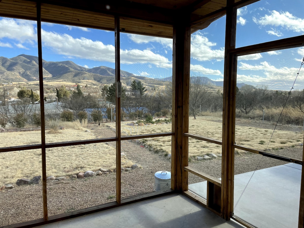 A spacious sunroom offers expansive floor-to-ceiling windows that frame sweeping views of the mountains and landscape. The area features wooden beams and a concrete floor, creating a bright space that connects indoor and outdoor environments.