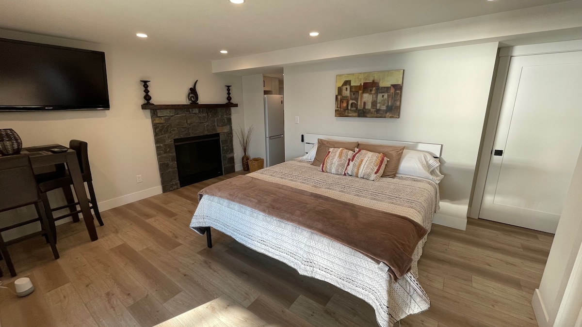 A spacious studio suite features a comfortable bed with neutral bedding, positioned in a well-lit room. A modern fireplace is set against a stone wall, while a Smart TV is mounted above. A dining area with two chairs occupies one corner of the room.