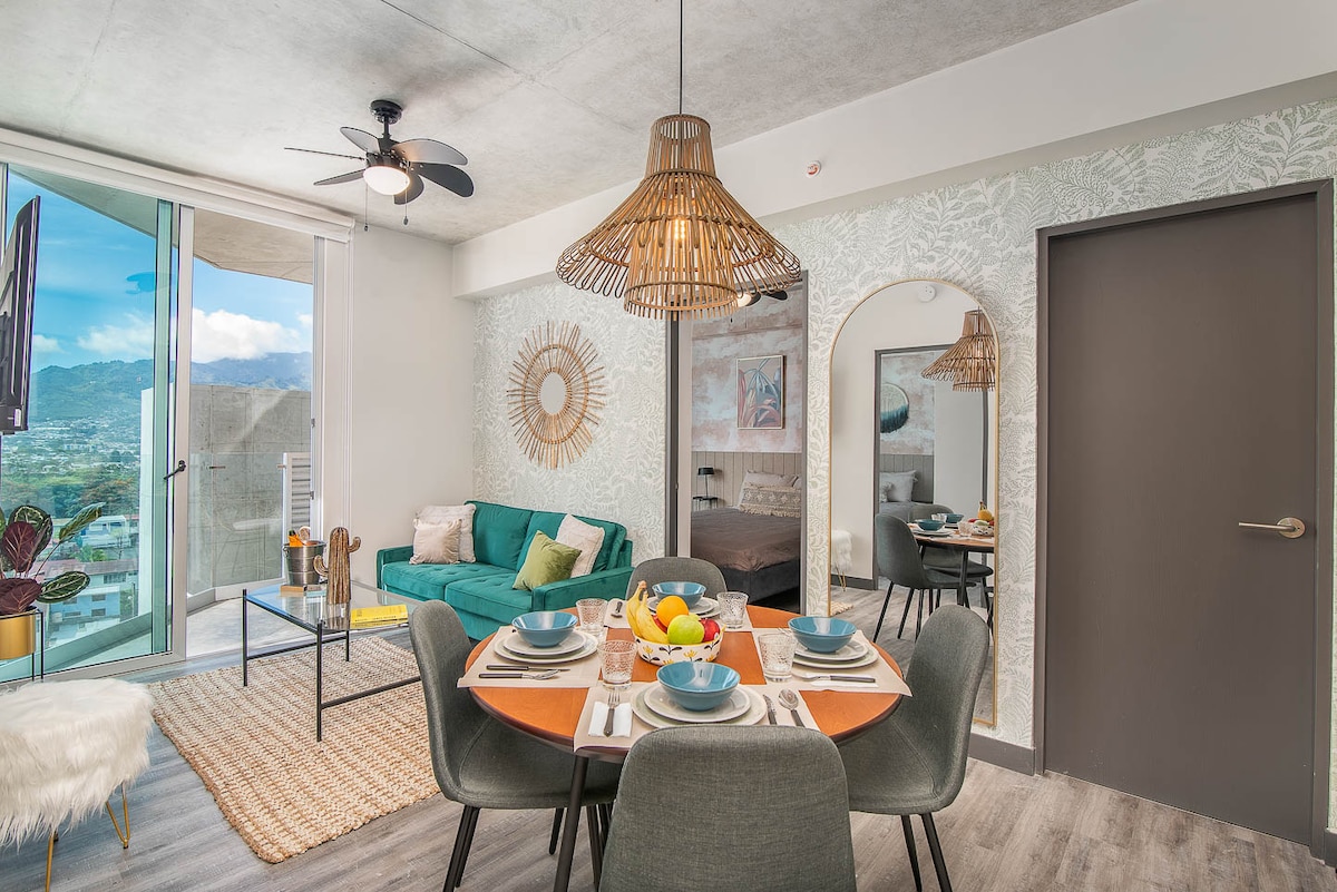 A bright and airy living space features a green sofa and a dining area with a round table set for four. Large windows allow natural light to fill the room, showcasing views of the surrounding landscape. Beautiful wall decor and a woven light fixture add to the charm.