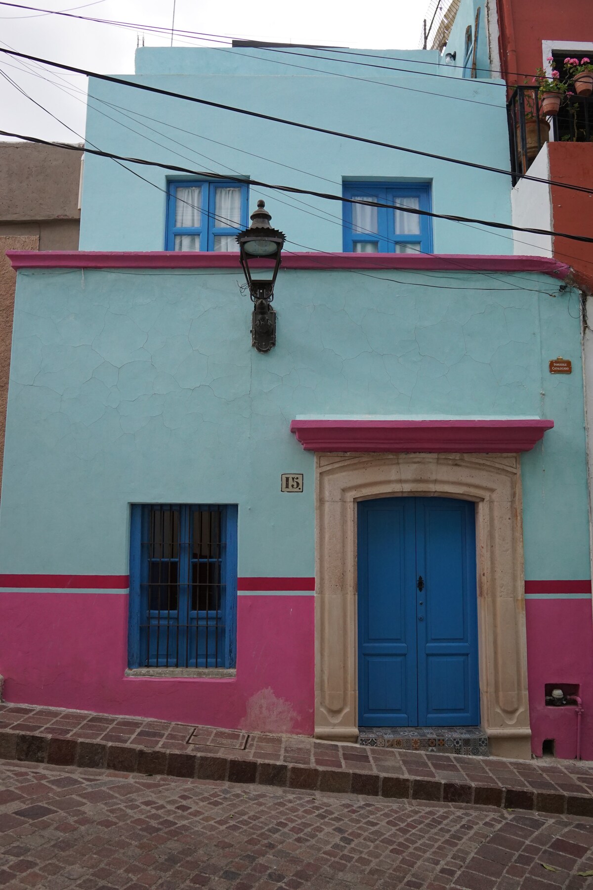 A vibrant exterior of a building is displayed, featuring a combination of blue, pink, and turquoise paint. The entrance is highlighted by a blue door and a decorative archway, while window bars add character to the façade. The area is lined with charming cobblestones.