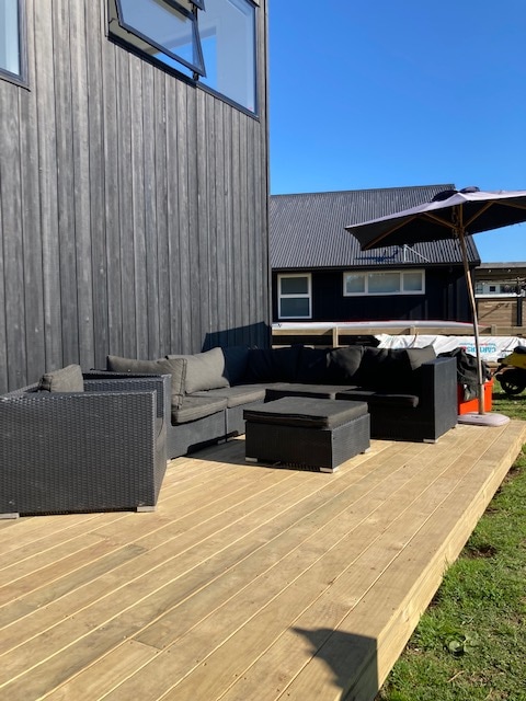 An outdoor deck area features a dark wicker sectional sofa and a matching coffee table. A large umbrella provides shade, while the backdrop includes two modern structures with a clear blue sky above. The deck's wooden flooring complements the contemporary aesthetic.