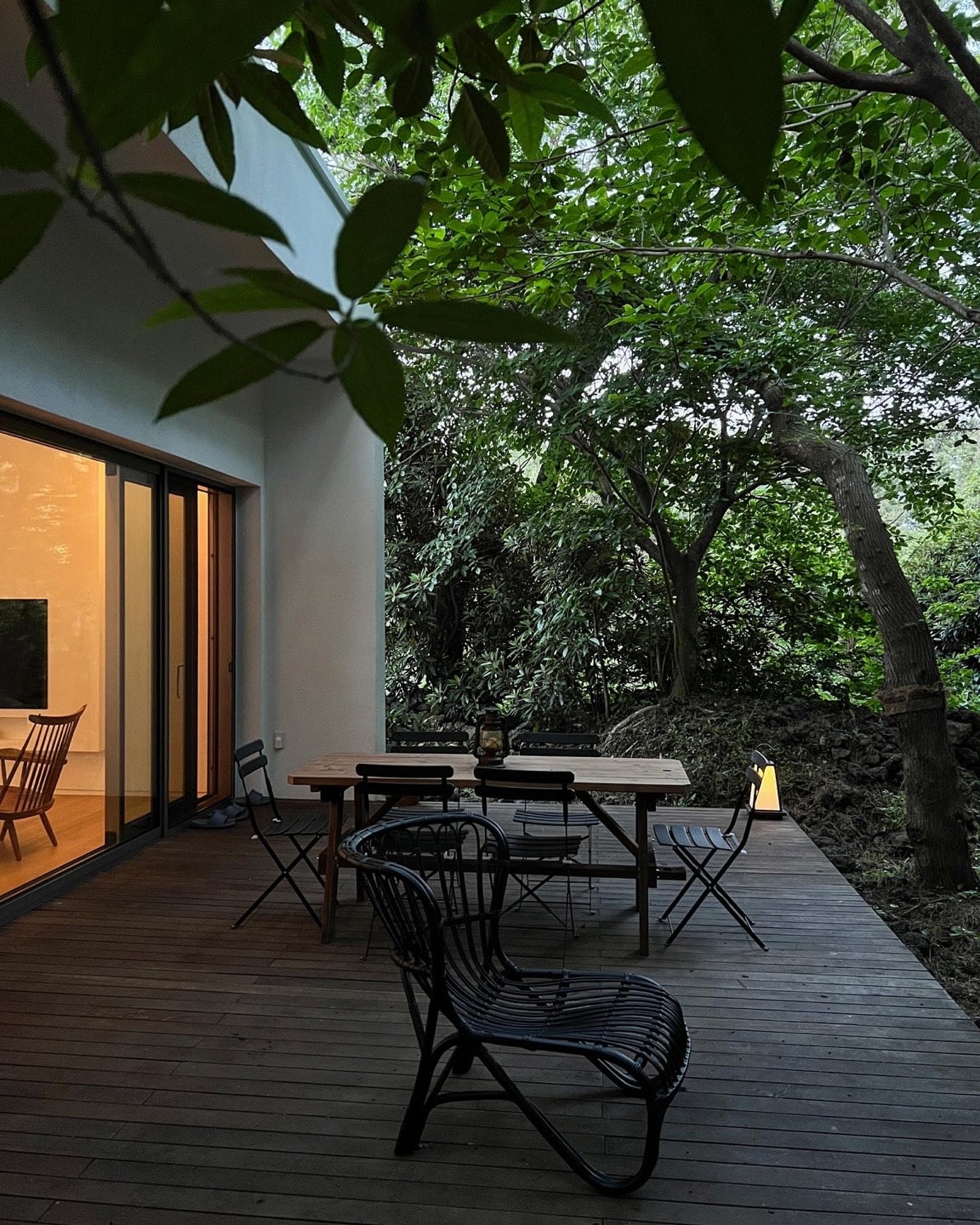 A wooden deck adorned with a table and several chairs is surrounded by lush greenery. Natural light filters through the leaves, creating a serene atmosphere. The adjacent glass doors provide a glimpse into the interior space, enhancing the connection with nature.