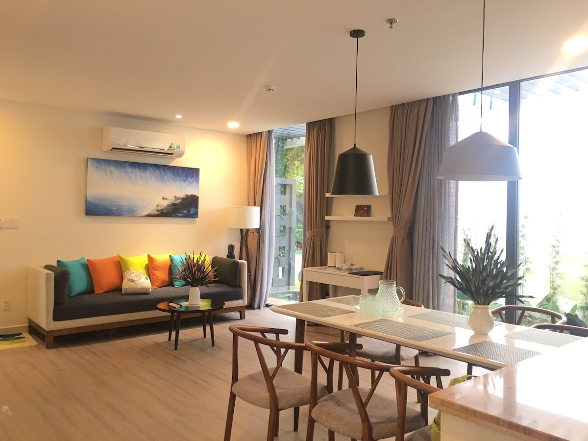 A modern living space is displayed, featuring a comfortable sofa adorned with colorful pillows. A dining table with elegant chairs is positioned nearby, complemented by stylish pendant lights. Large windows invite natural light, providing views of greenery outside.