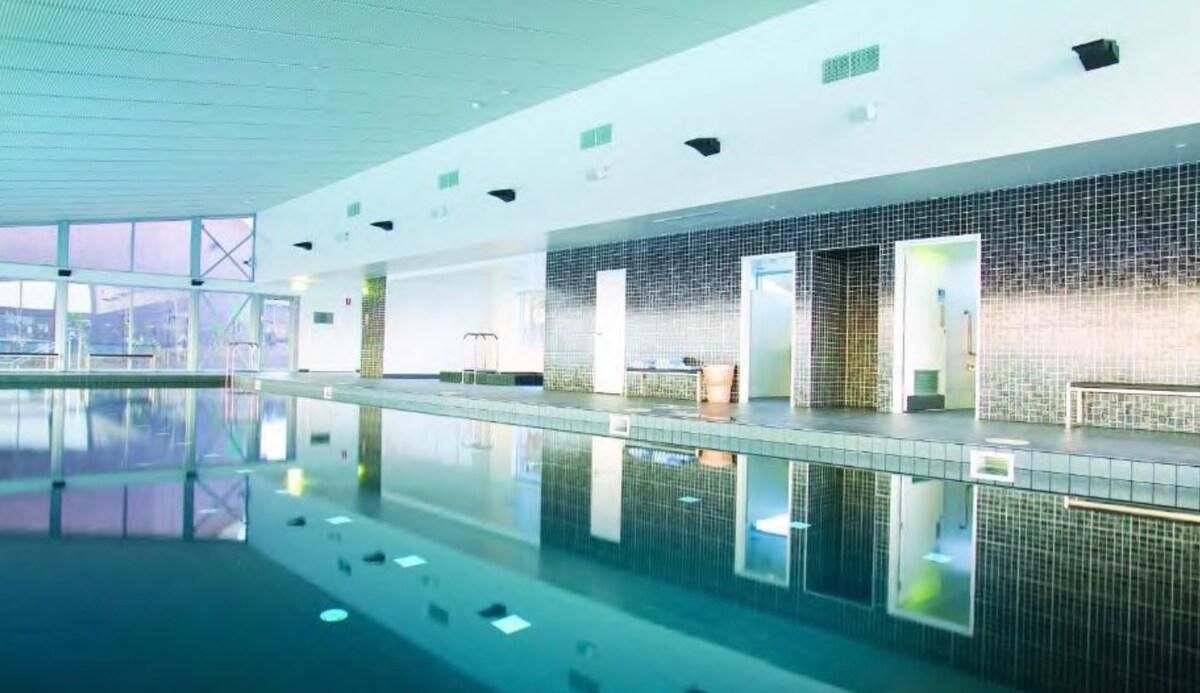 An indoor pool area features a calming water surface reflecting the surrounding textured tiles and softly lit recessed lighting. Multiple entry points to adjoining spaces can be seen along the smooth walls, creating an open and inviting atmosphere.