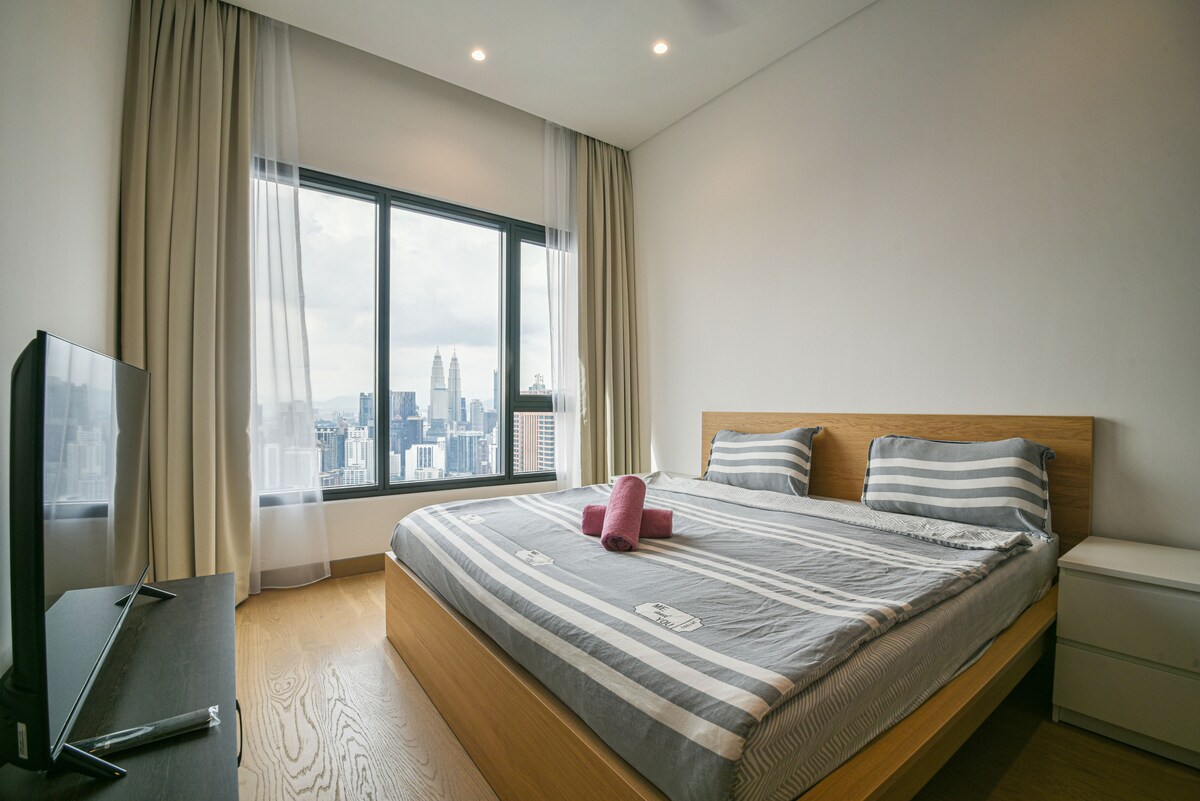A spacious bedroom features a large bed with striped linens, flanked by windows showcasing a city skyline. Light curtains allow natural light to fill the room, while a television is positioned on a dark-hued cabinet, creating a contemporary and comfortable environment.