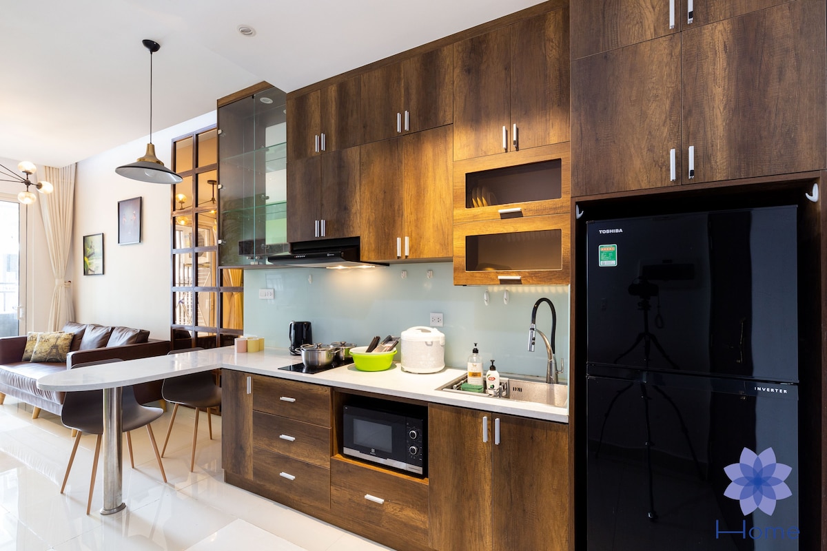 A modern kitchen is presented with wooden cabinetry and a sleek countertop. Stainless steel appliances are visible, including a microwave and a refrigerator. Natural light filters through a nearby window, illuminating the space. An inviting dining area features a marble table and comfortable seating.