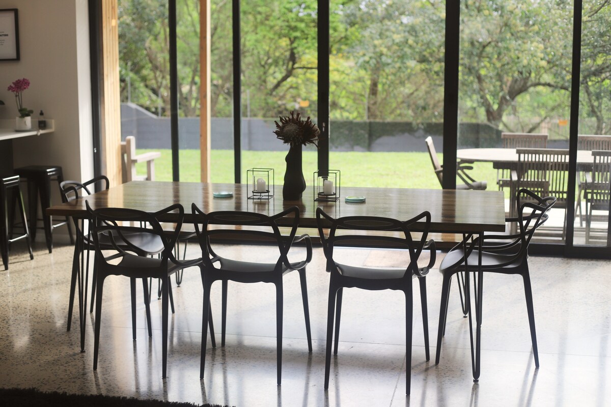 A modern dining area features a large wooden table surrounded by six black chairs. Floor-to-ceiling glass doors invite natural light and provide a view of the green garden outside. Decorative elements include a vase with flowers and candles on the table.