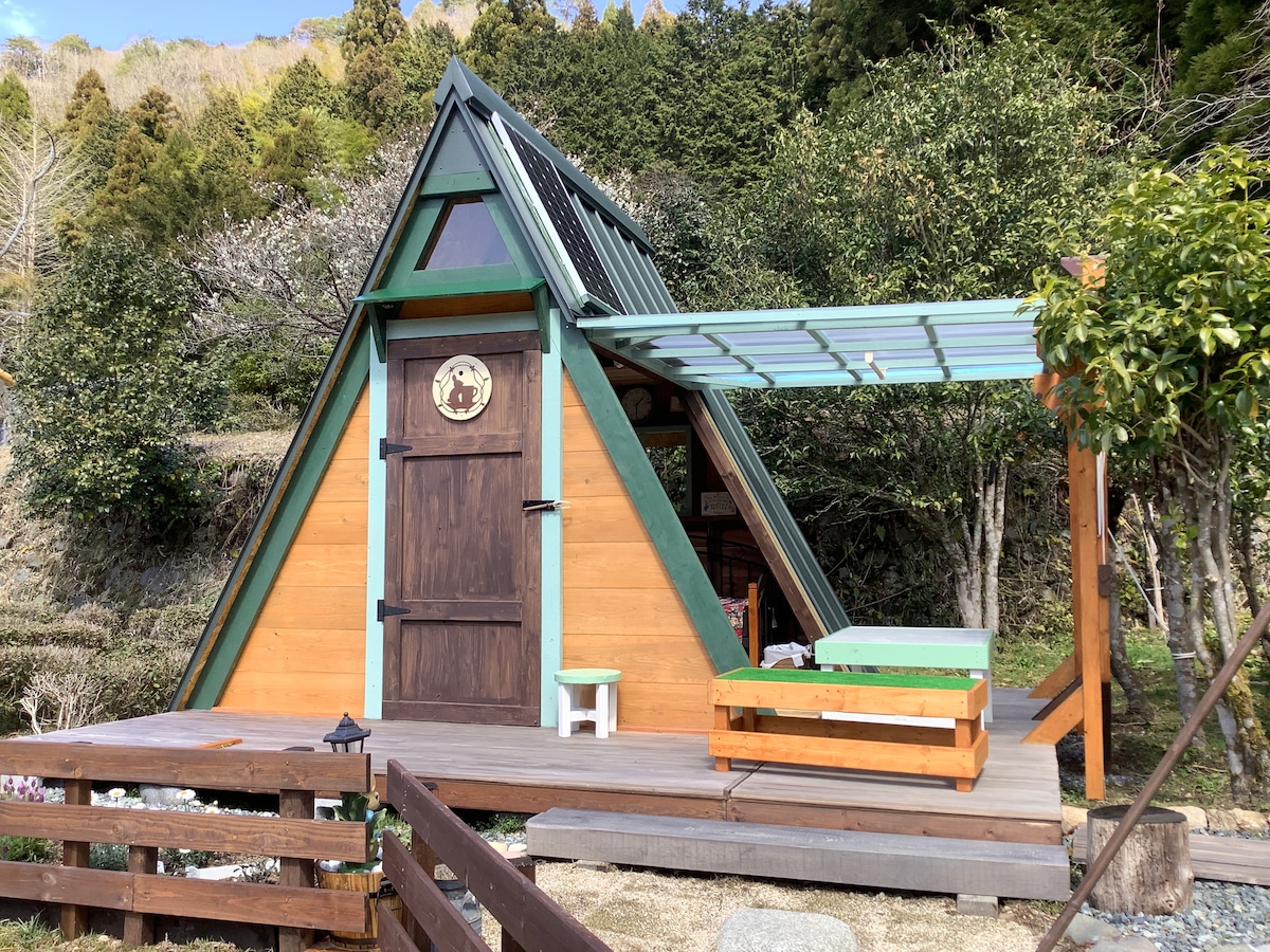An A-frame cottage is featured, showcasing natural wood siding with green accents. A covered area is provided on the porch, with a small seating space visible. Surrounding greenery adds to the tranquil setting, creating an inviting atmosphere for outdoor experiences.